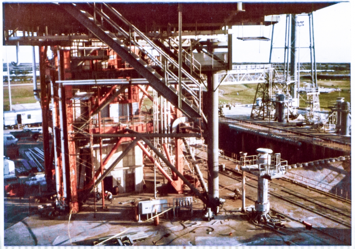 Beneath the looming darkness of the RSS which rises far into the sky above you, a photograph showing the view from the stair tower on Column Line 7 of the Rotating Service Structure at Space Shuttle Launch Complex 39-B, Kennedy Space Center, Florida, looking back toward the bottom of the RSS Hinge Column and Fixed Service Structure, and the pad itself below it and surrounding it, can only hint at the true immensity, grandeur, power, and symmetry of what you’re seeing. Visible in this image, among much, very much, more, you can see the Flame Trench, MLP Mount Pedestals, Sound Suppression Water Spray Headers and the bottom of the SSW Water Tower, the North Piping Bridge, the Crawlerway Tracks on the Pad Deck, MLP Access Stairs protruding down into empty space beneath the RSS, Hinge Column Struts, a workman bent over his work in front of the FSS Elevator Door, and the arrayed elements of an active construction site visible in all directions. It was a marvelous place, at a marvelous time to be there, and I shall never forget just how fortunate I was to be able to participate in it.